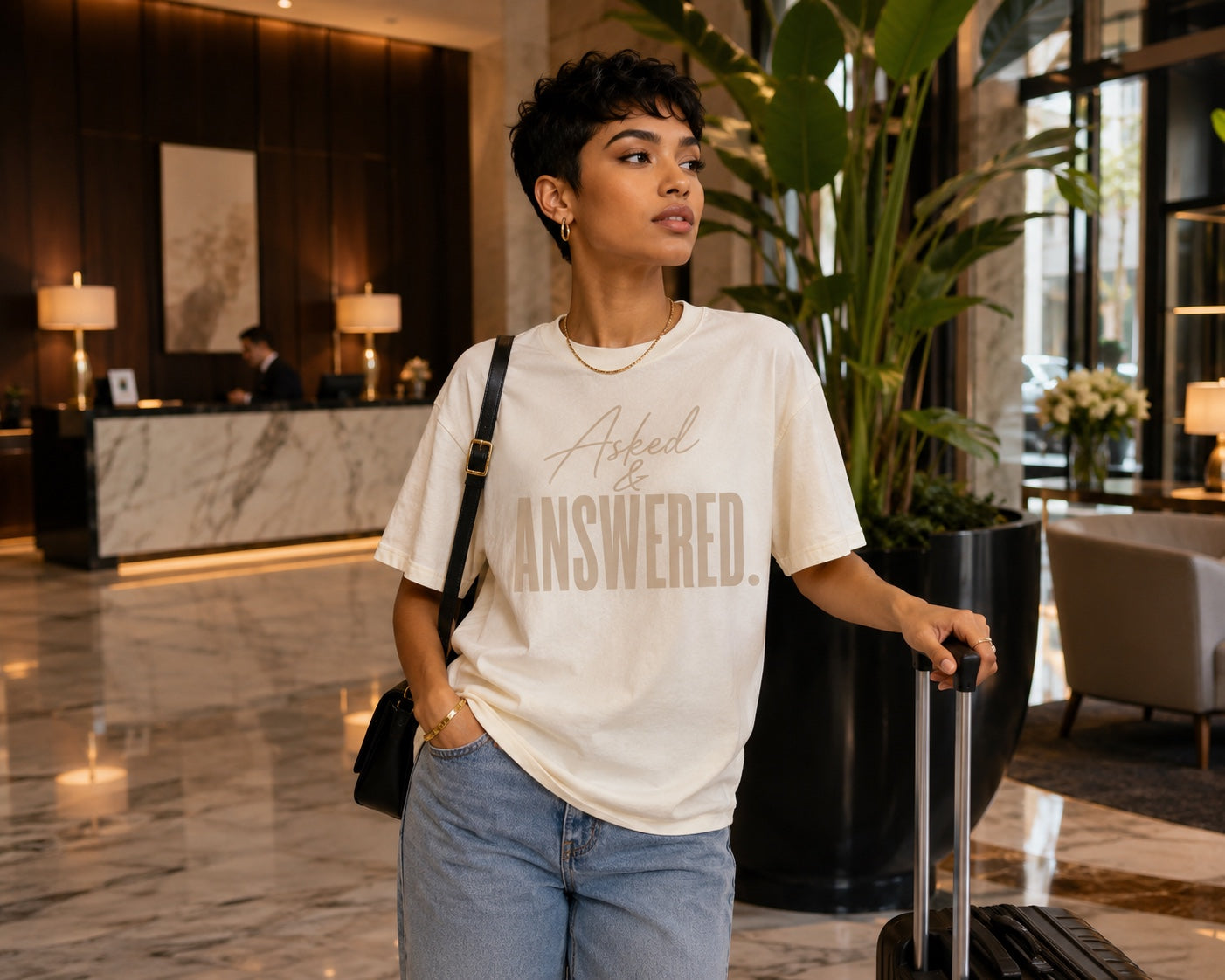 Woman wearing the Asked & Answered Comfort Colors Ivory tee with jeans and luggage in a luxury hotel lobby.