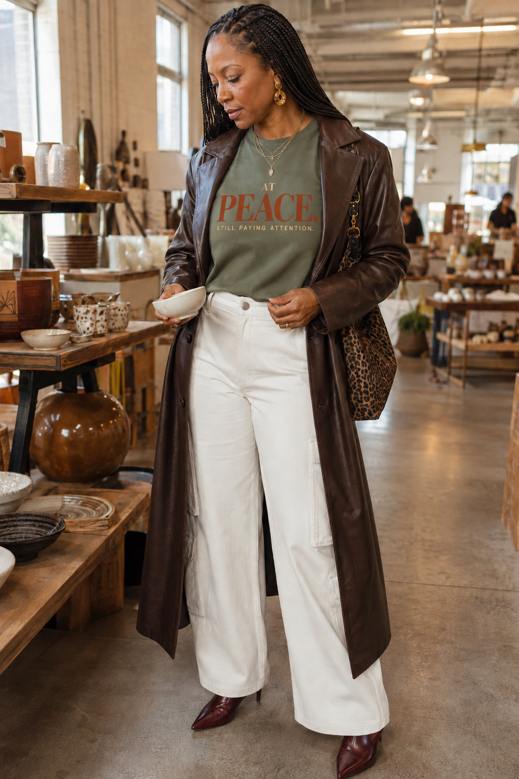 Woman wearing the At Peace Still Paying Attention Comfort Colors Moss statement tee with a brown leather trench and white trousers.
