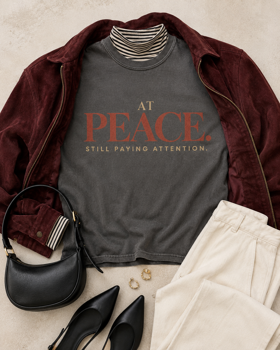 Styled flat lay of the At Peace Still Paying Attention Comfort Colors Pepper Gray statement tee with an oxblood jacket, ivory trousers, black accessories, and gold jewelry.