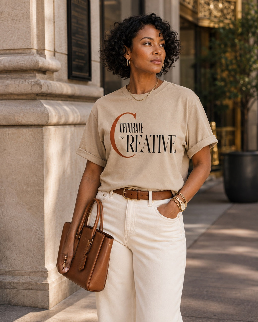 Woman wearing the Corporate to Creative statement tee in Comfort Colors Sandstone with cream denim and a structured leather bag