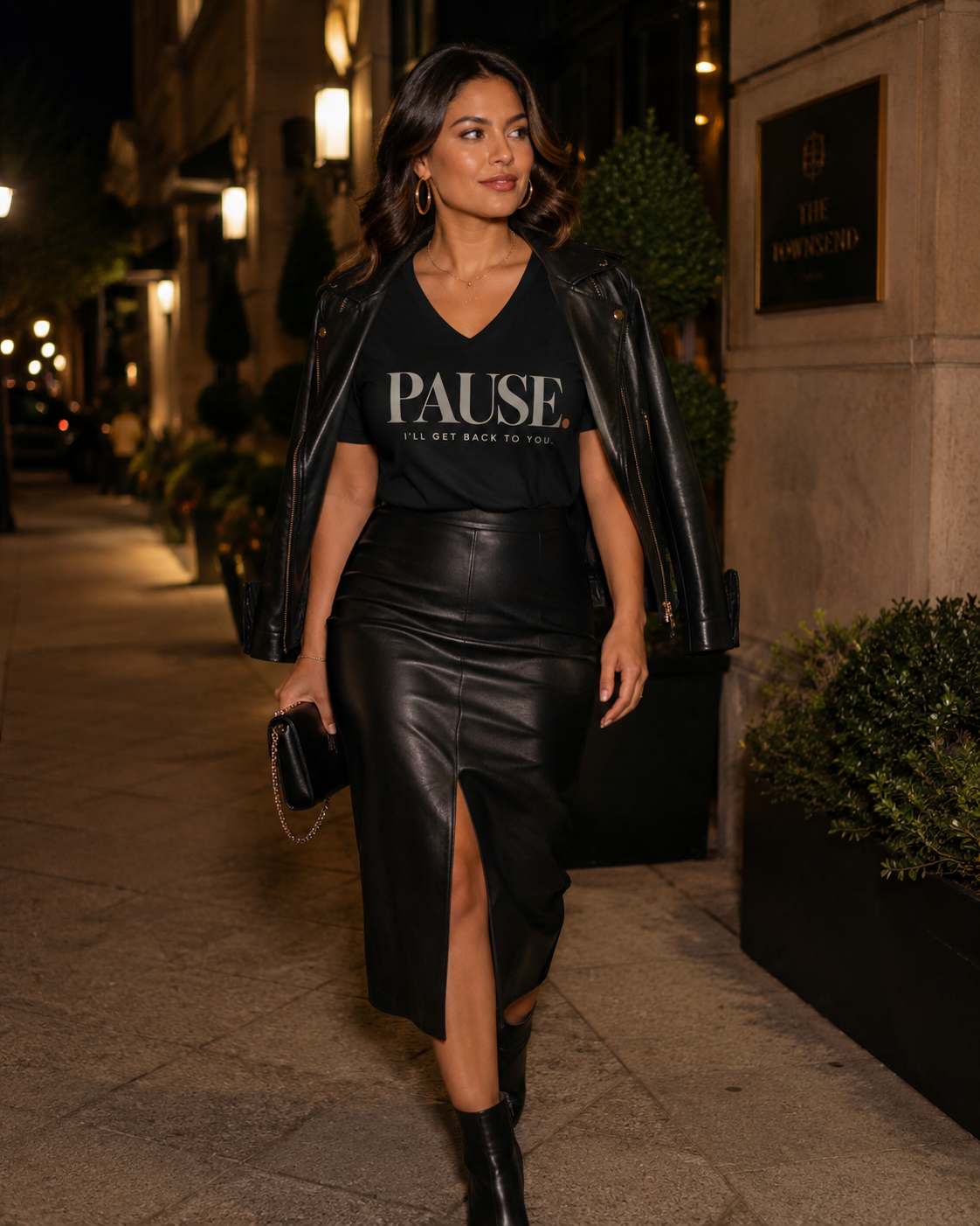 Woman wearing the PAUSE. I'll Get Back to You. women's V-neck statement tee in Black with a leather jacket, leather midi skirt, and clutch