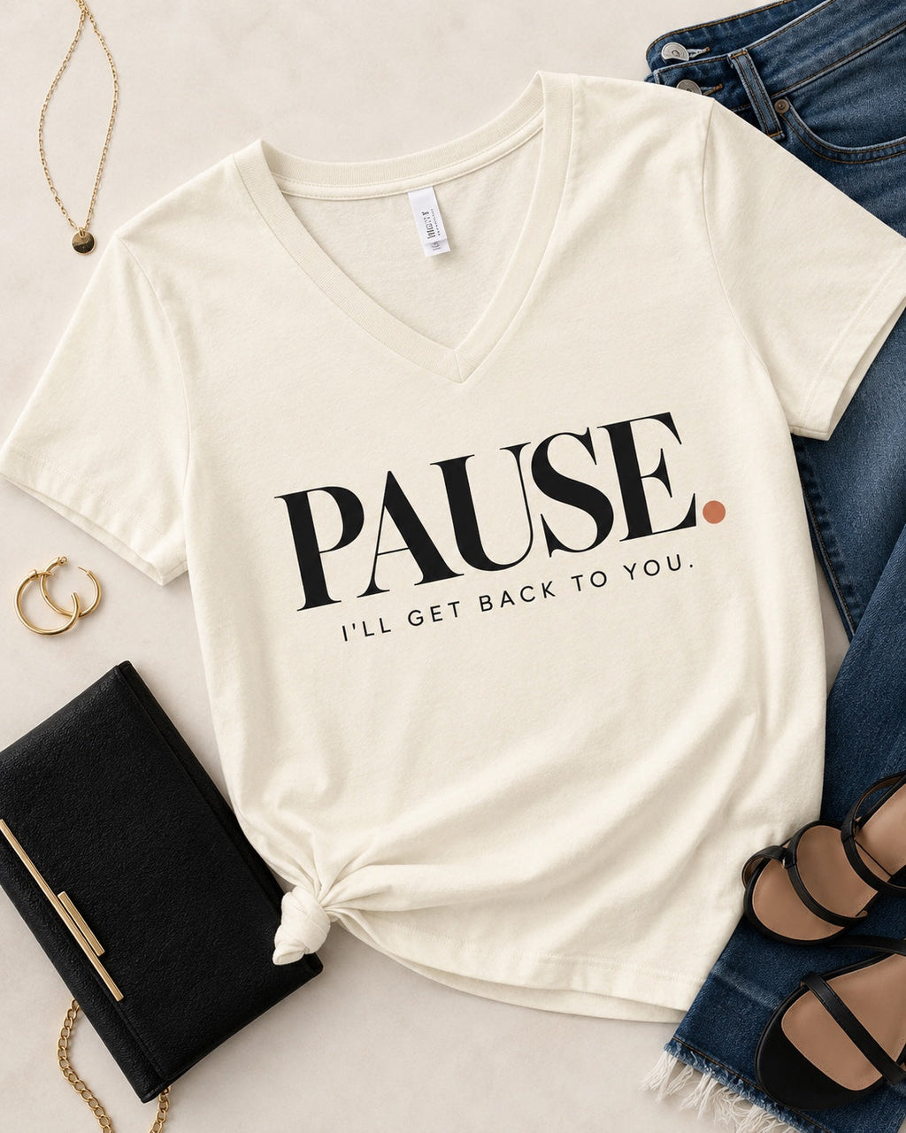 Styled flat lay of the PAUSE. I'll Get Back to You. women's V-neck statement tee in Natural with denim, black clutch, gold jewelry, and black heels