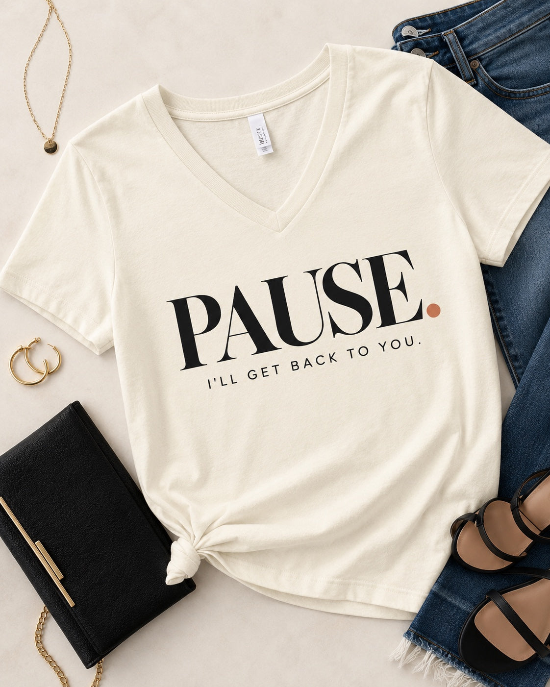Styled flat lay of the PAUSE. I'll Get Back to You. women's V-neck statement tee in Natural with denim, black clutch, gold jewelry, and black heels