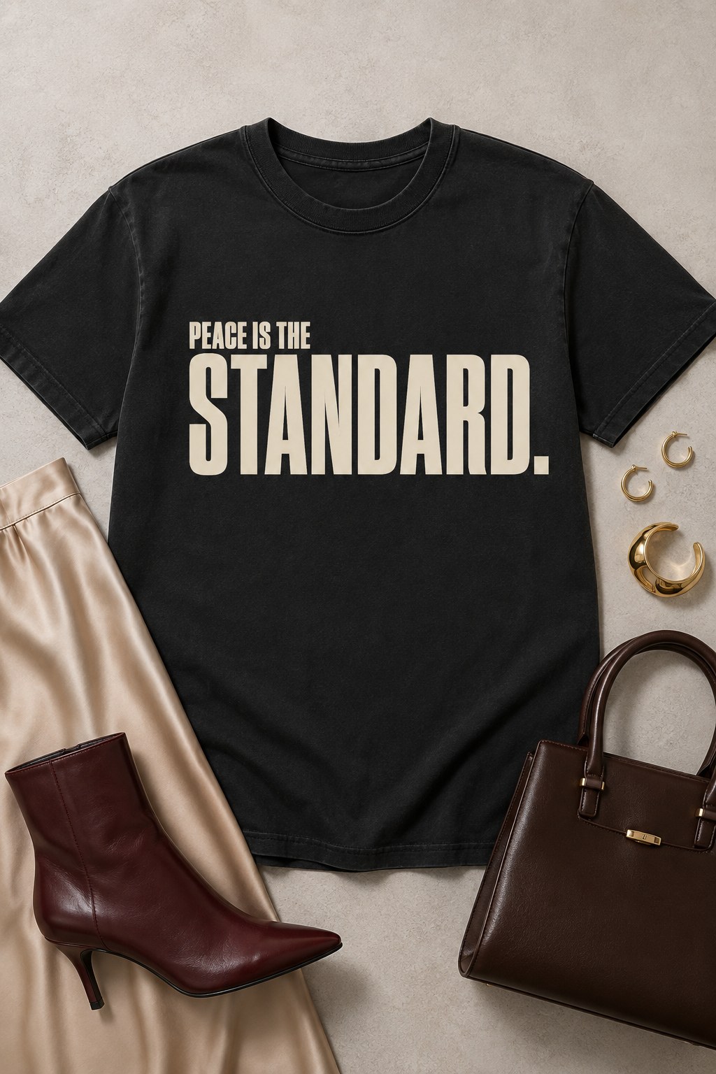 Styled flat lay of the Peace Is the Standard Comfort Colors Black statement tee with a champagne satin skirt, oxblood boot, dark brown handbag, and gold jewelry.